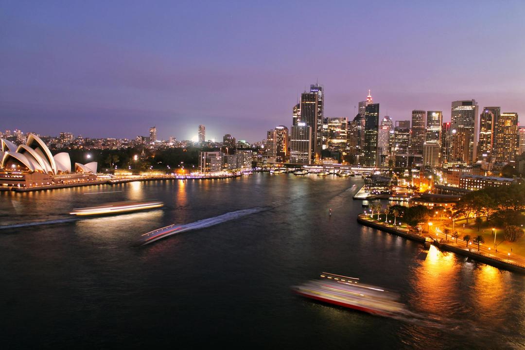 The waterfront in Sydney, Australia, depicting the opera house and some boats on the water at sunset, in front of the Sydney skyline.