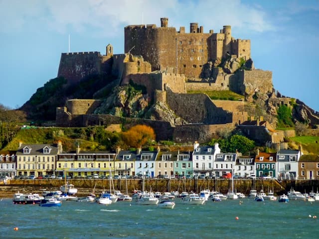 Colourful houses in front of large stone castle, next to sea with docked sailing boats.